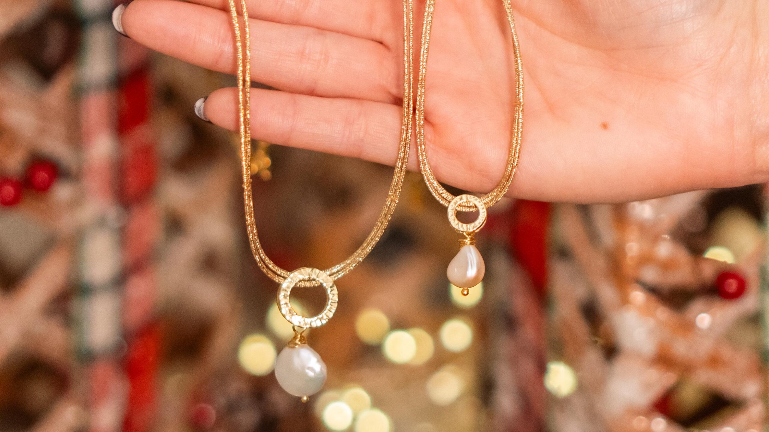 Una mano elegante tiene due collane d'oro con pendenti di perle, brillando davanti a un albero di Natale sfocato.